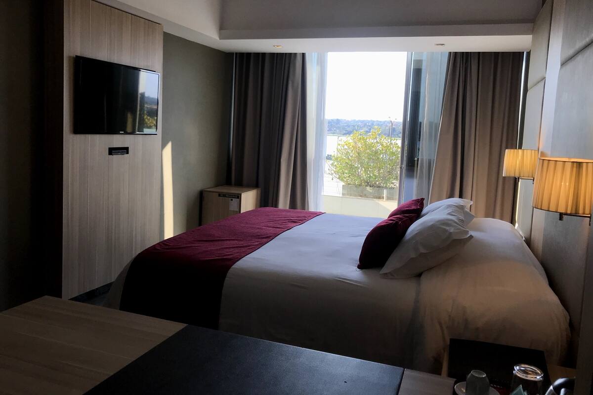 A hotel room with a bed, a desk, a television, a window with curtains, and a view of the outdoors.