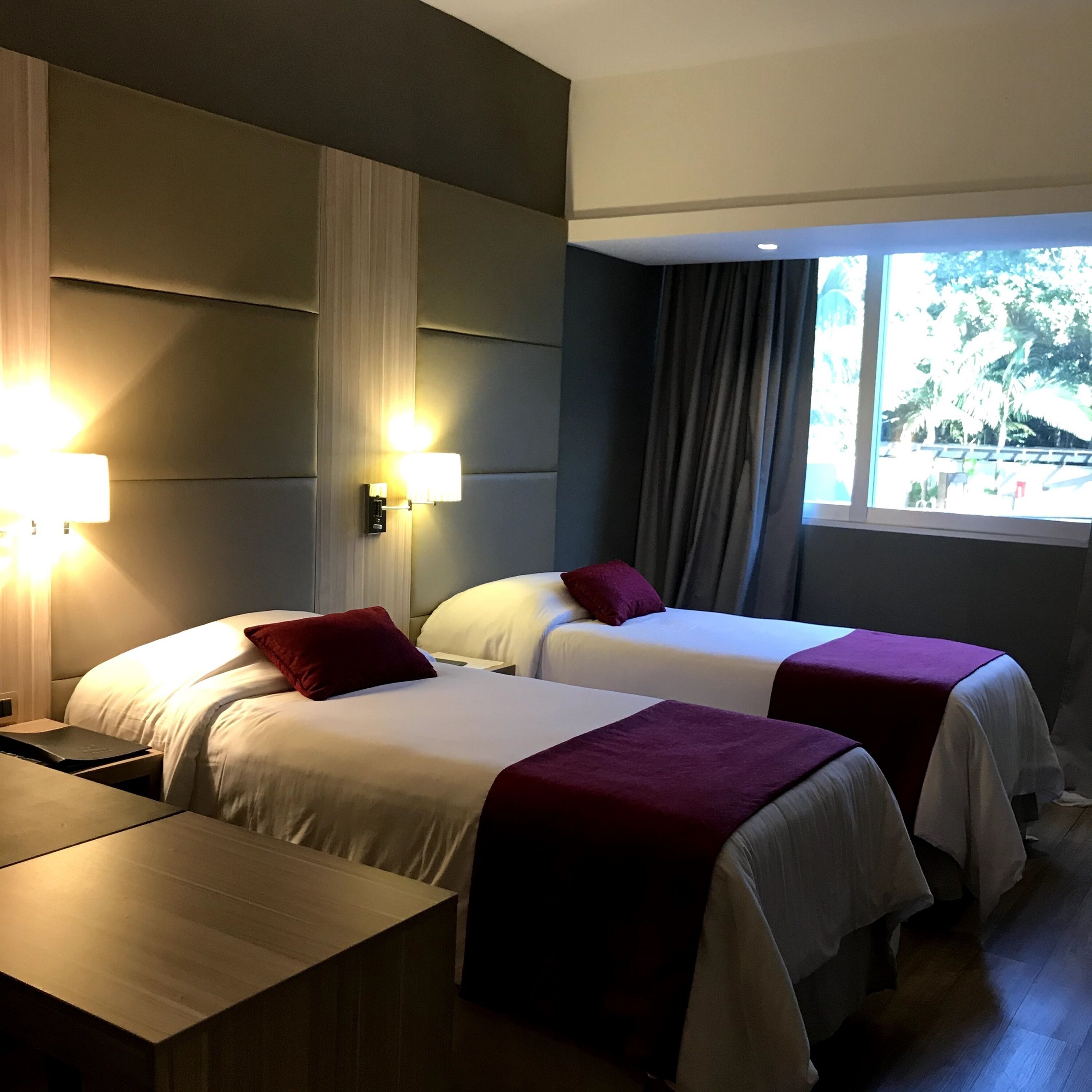 A hotel room with two beds, a desk, and a window with a view of trees.