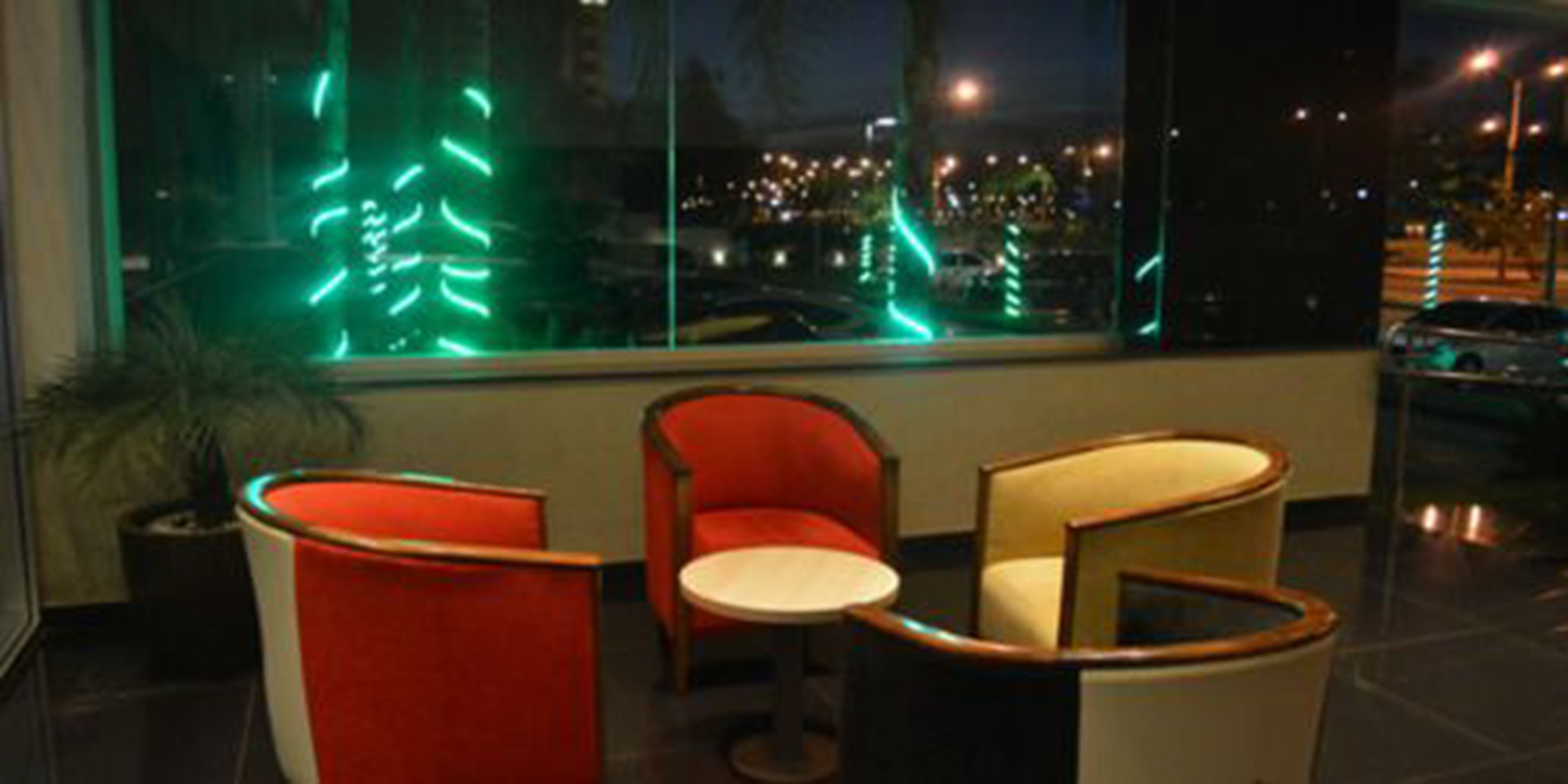 Un interior moderno con dos sillones rojos y una mesita, y un ventanal con vista a un paisaje urbano nocturno.