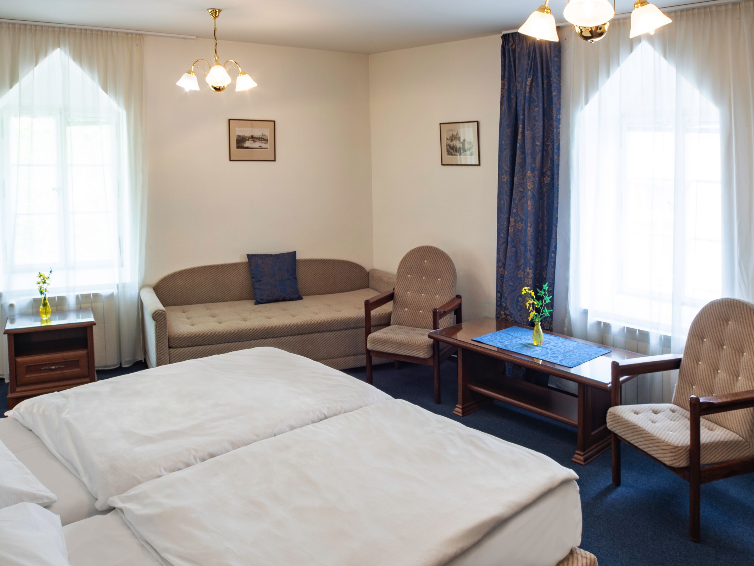 A hotel room with a large bed, a sofa, two armchairs, a small table with a vase, and two windows with curtains.