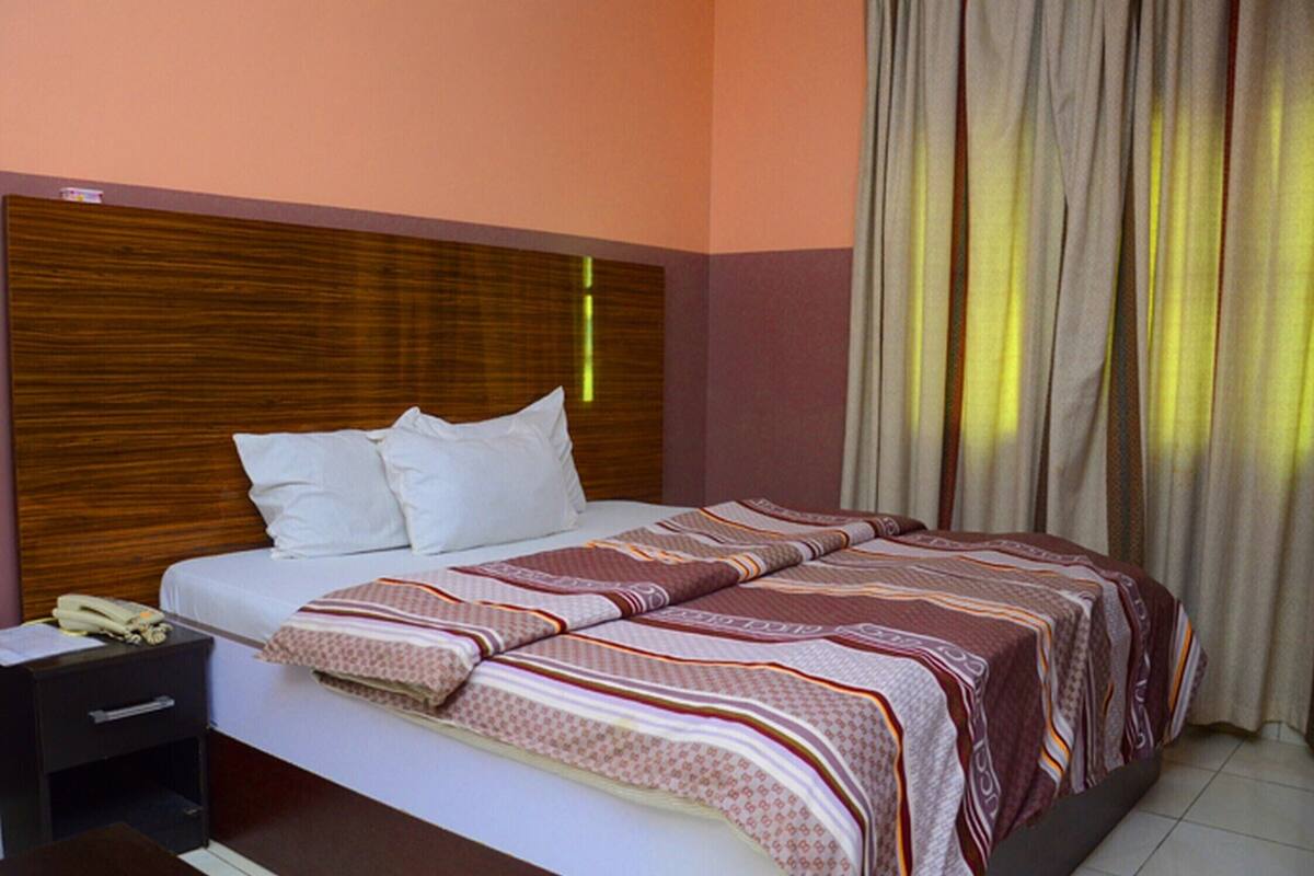 A hotel room with a wooden headboard, a bed with a patterned bedspread, a nightstand with a telephone, and a window with curtains.