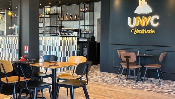 Angolo bar con scaffali pieni di bottiglie, un bancone e un cartello che riporta 'UNYC Brasserie'.