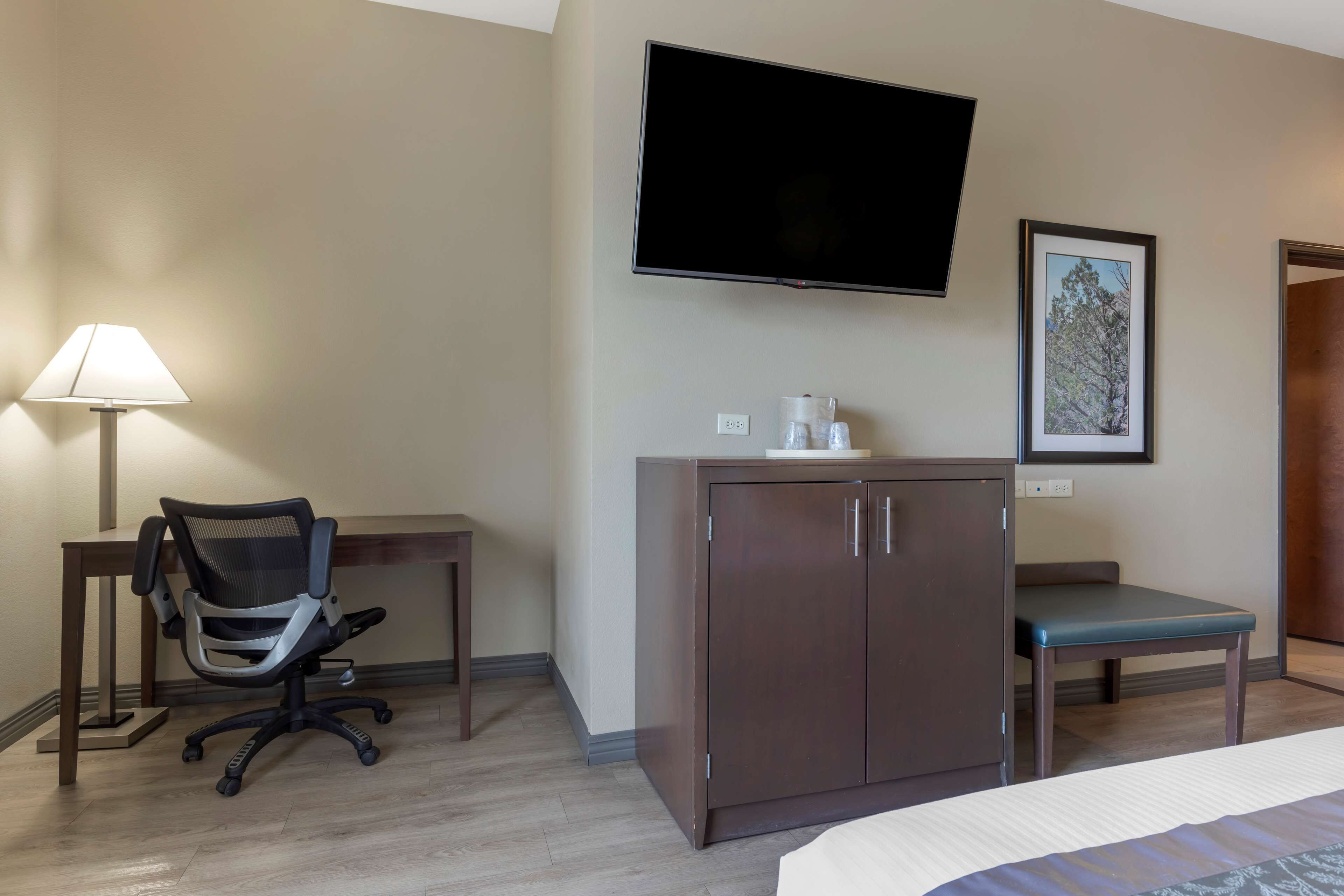 Una habitación de hotel con una cama, un escritorio con una silla, un televisor montado en la pared, un armario, un cuadro enmarcado y un banco.