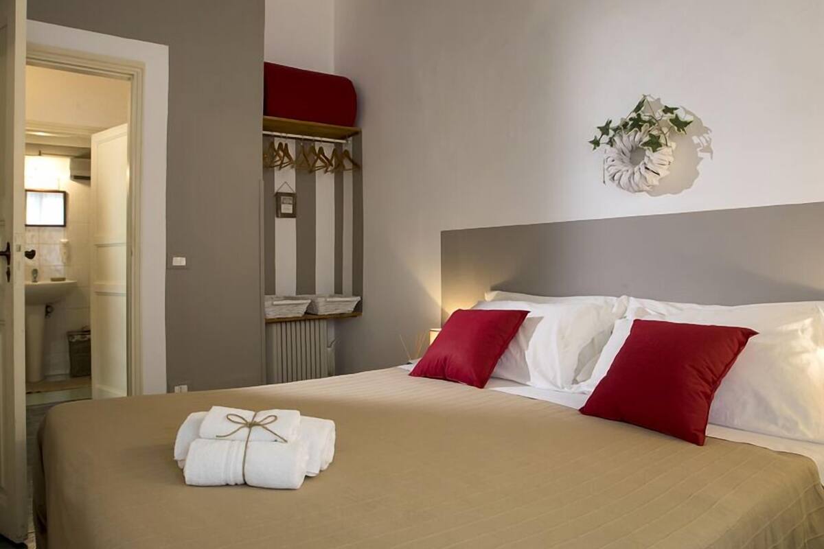 A hotel room with a bed, a red and white headboard, a wall-mounted plant, and a red towel on the bed.