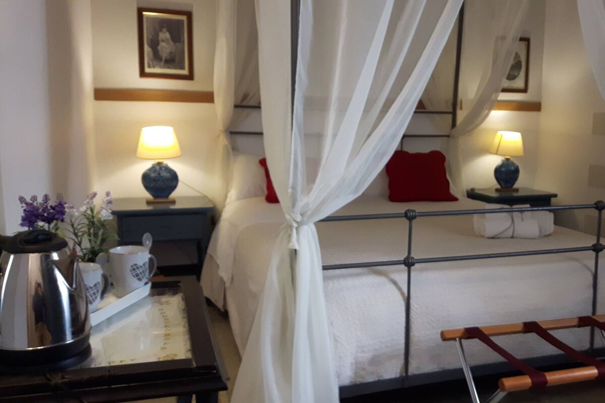 A hotel room with a bed, two bedside tables, a nightstand, a lamp, a vase, and a small table with a kettle and cups.