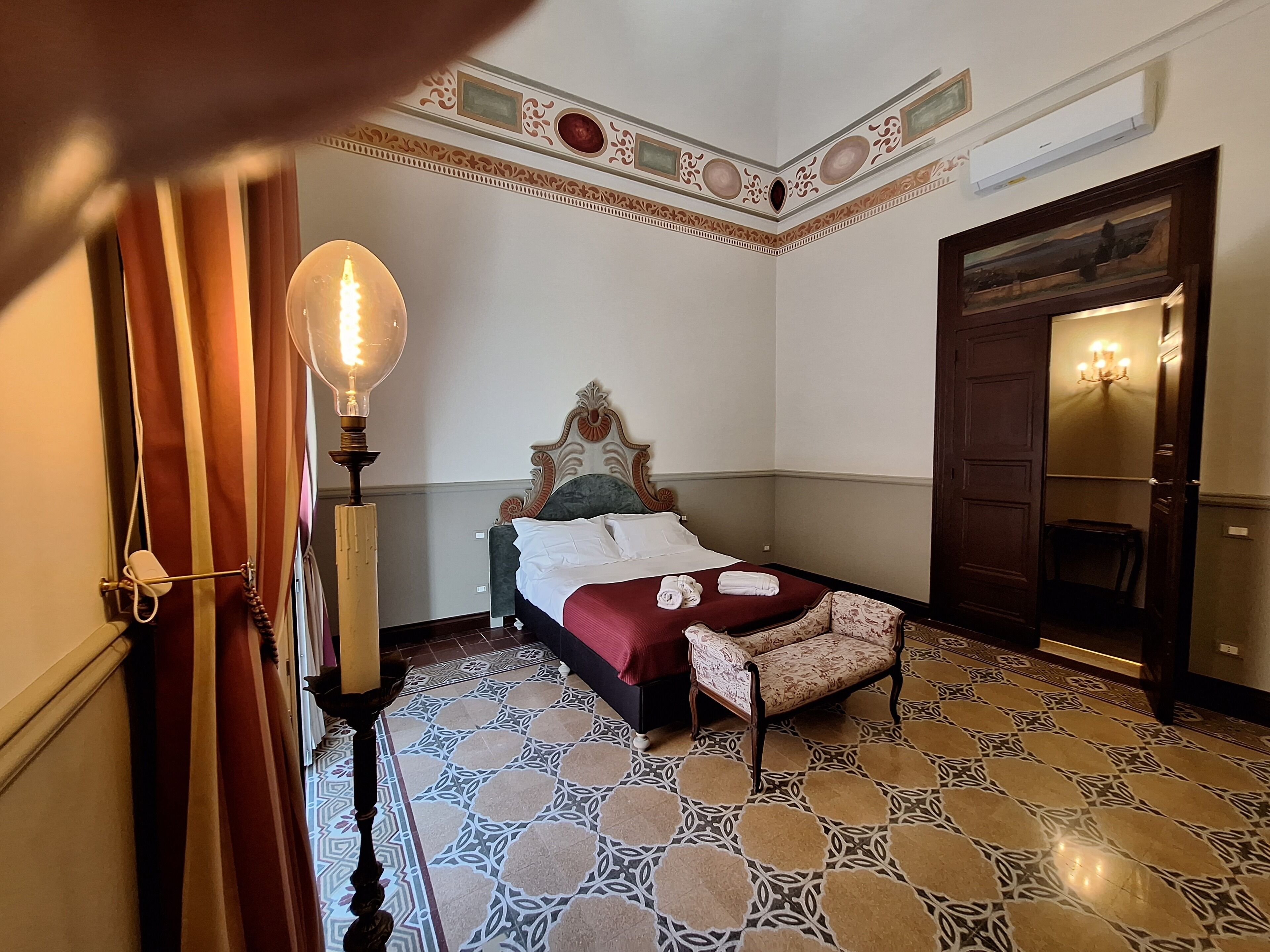 A hotel room with a bed, a nightstand, a lamp, a patterned floor, and a door.