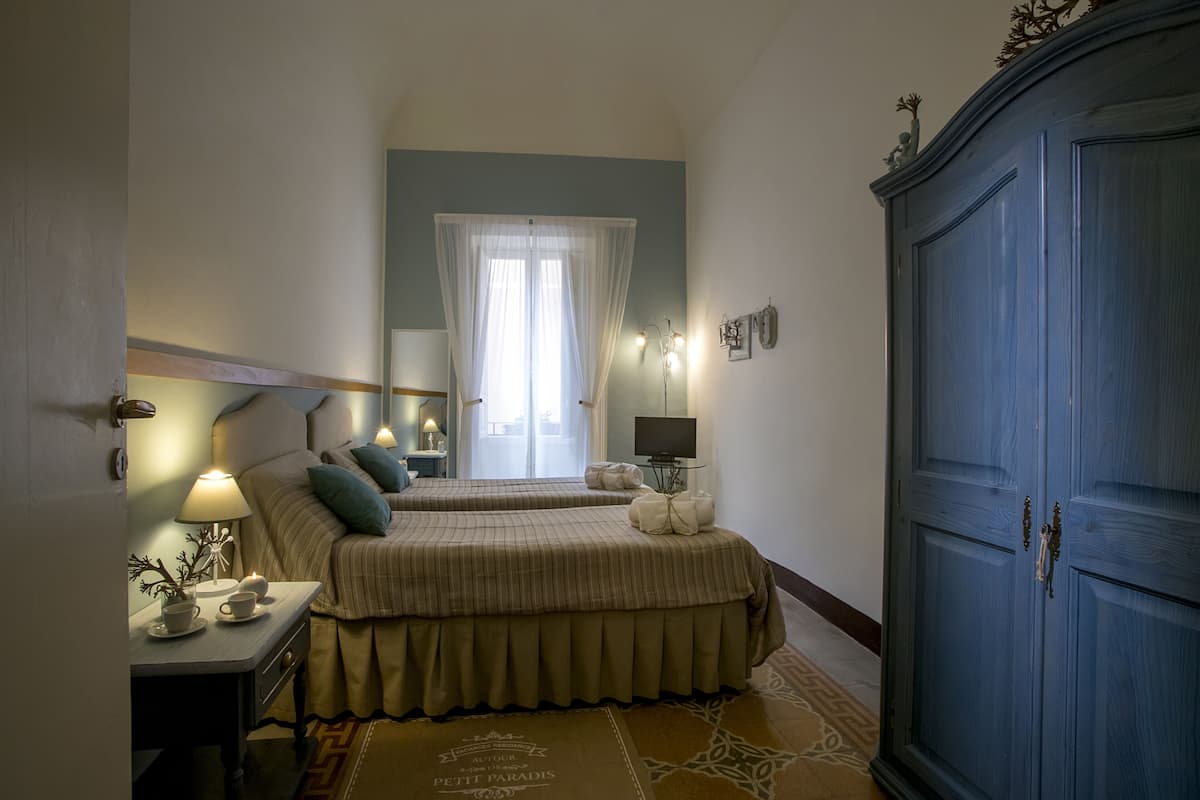 A hotel room with two beds, a wardrobe, a bedside table with a lamp, a small table with a plant, and a window with sheer curtains.