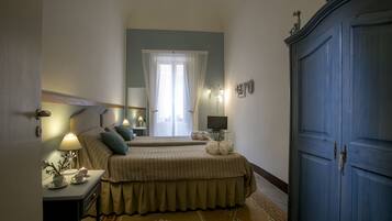A hotel room with two beds, a wardrobe, a bedside table with a lamp, a small table with a plant, and a window with sheer curtains.