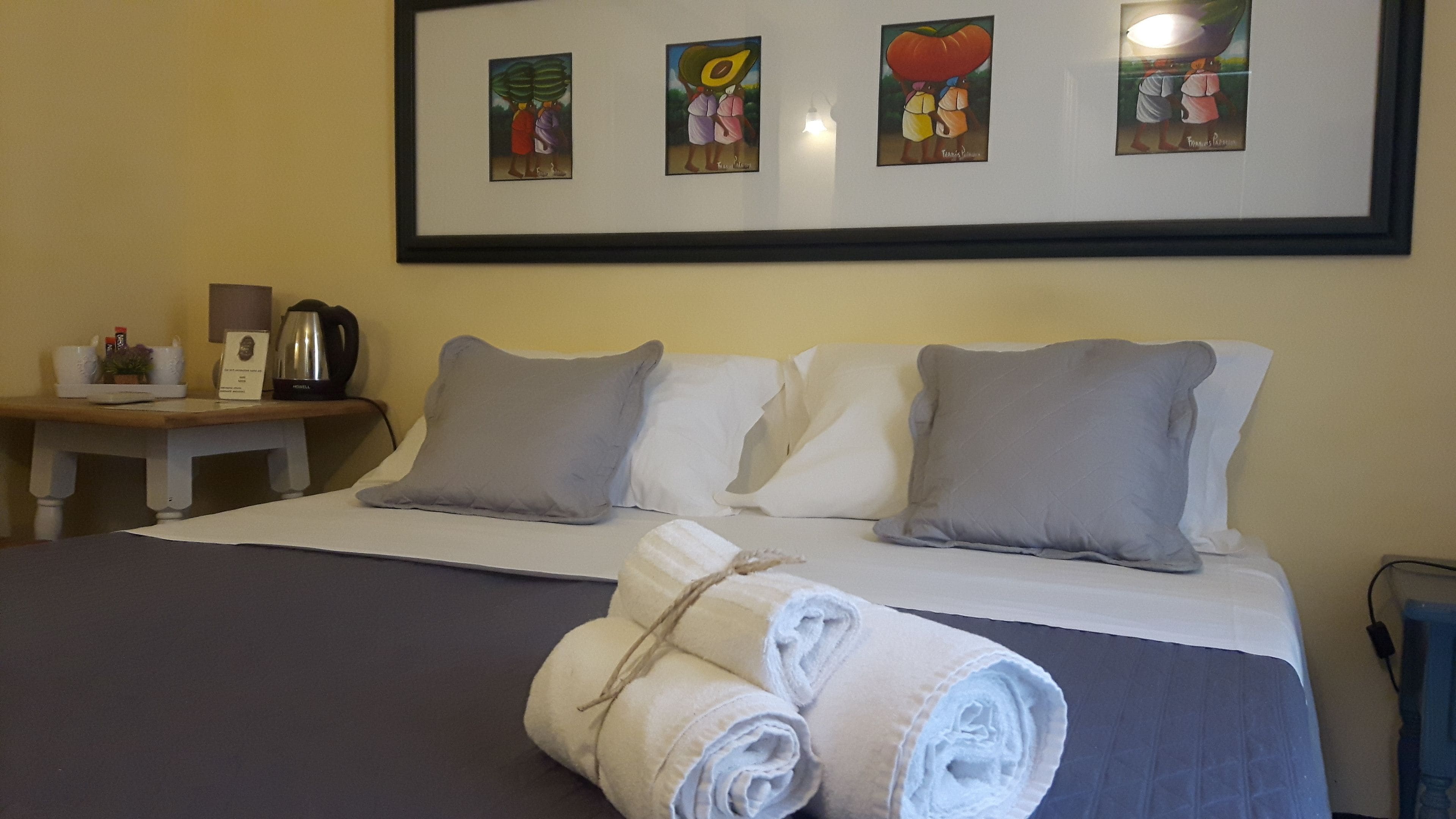 Una cama bien hecha con almohadas grises y blancas, una mesita de noche con una tetera y una planta pequeña, y una obra de arte enmarcada sobre la cama.