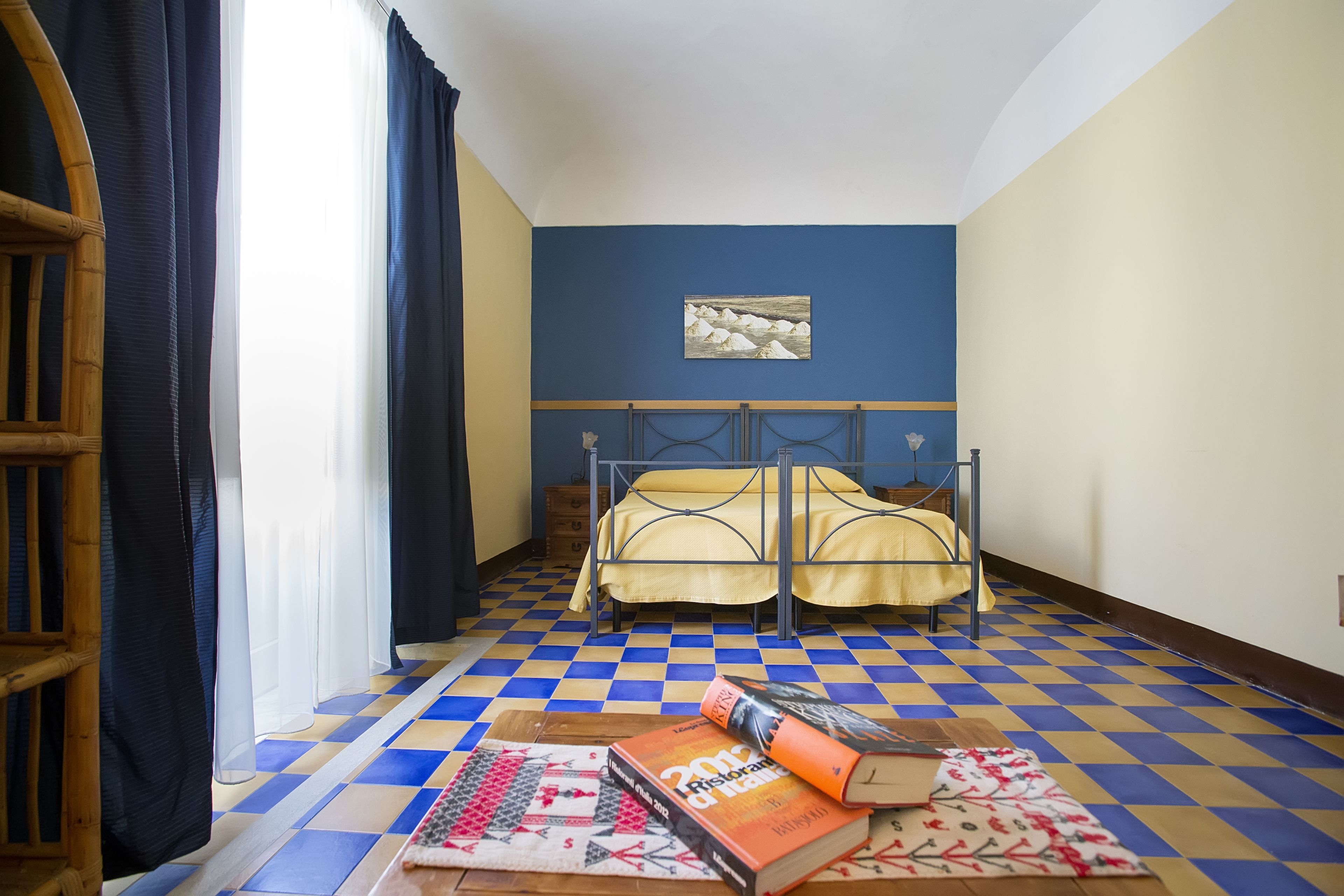A room with a checkered floor, two beds with yellow bedding, a wooden chair, and a window with sheer curtains.