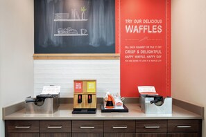 A waffle station with two waffle batter dispensers and a sign advertising waffles. - Hampton Inn & Suites Minneapolis / Downtown (Minneapolis)