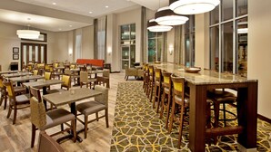 Ein Hotelrestaurant mit mehreren Tischen und Stühlen, großen Fenstern und einem gemusterten Teppich.
