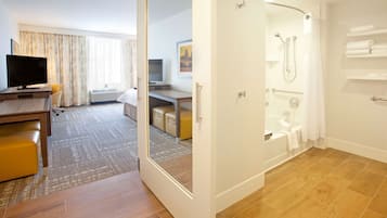 A hotel room with a wooden floor, a desk with a flat-screen TV, a sofa, a shower area, and a bathroom with a bathtub and towel rack.