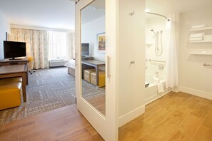A hotel room with a wooden floor, a desk with a flat-screen TV, a sofa, a shower area, and a bathroom with a bathtub and towel rack. - Hampton Inn & Suites Minneapolis / Downtown (Minneapolis)