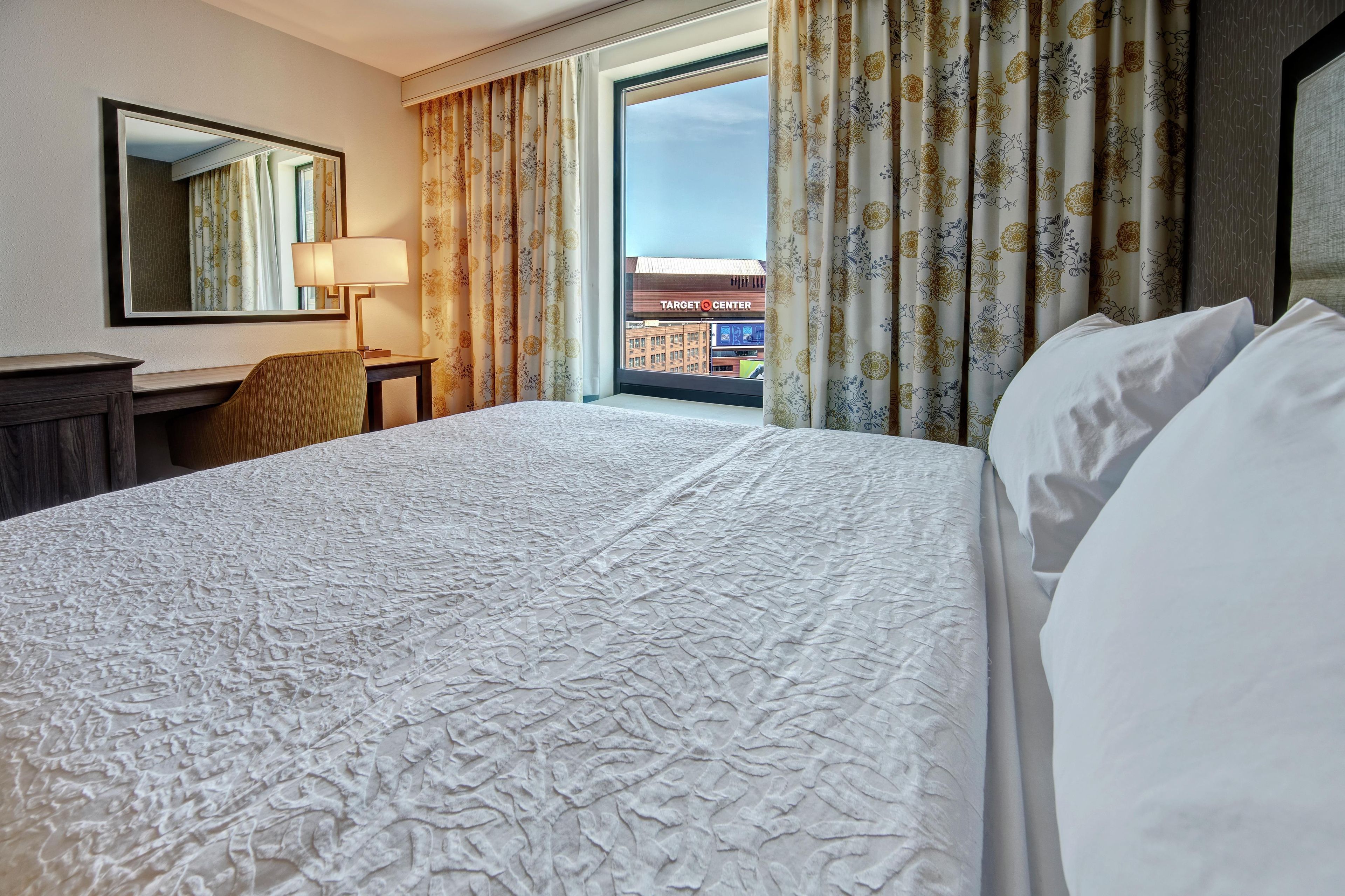 A hotel room with a large bed, a desk, a chair, a lamp, and a view of a cityscape through the window.