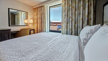 A hotel room with a large bed, a desk, a chair, a lamp, and a view of a cityscape through the window.
