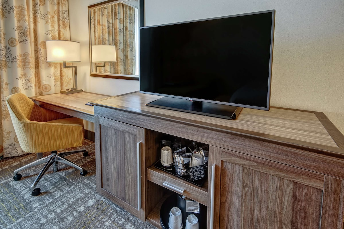 A hotel room with a flat-screen TV, wooden desk, cushioned chair, and a lamp.
