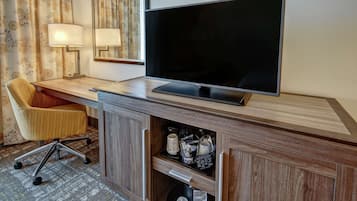 Quarto de hotel com televisão de tela plana, escrivaninha de madeira, cadeira estofada e luminária.