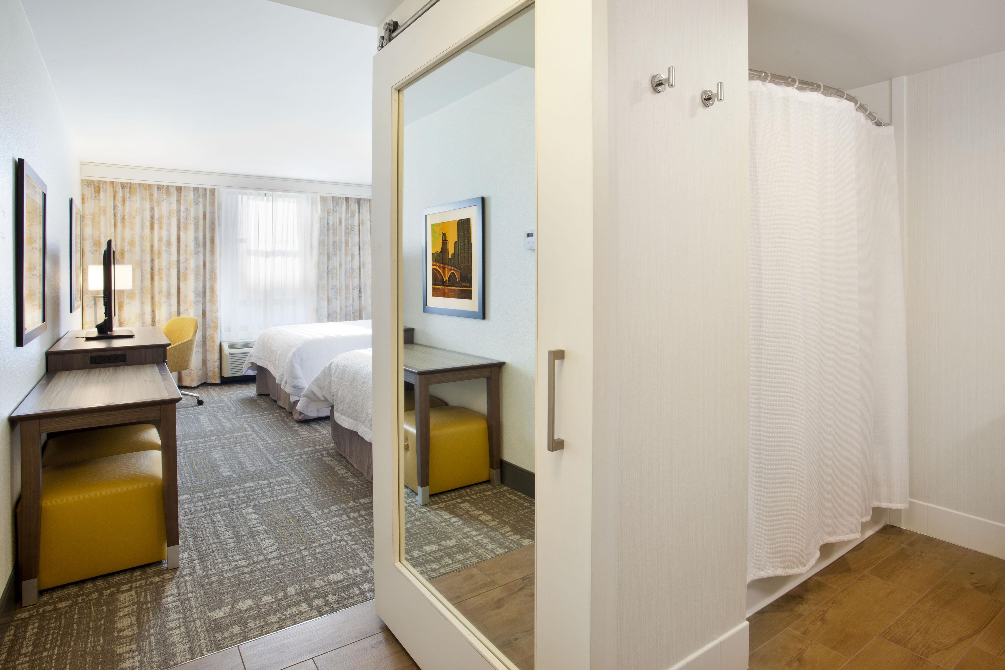 Habitación de hotel con cama, escritorio, silla, espejo y zona de ducha.