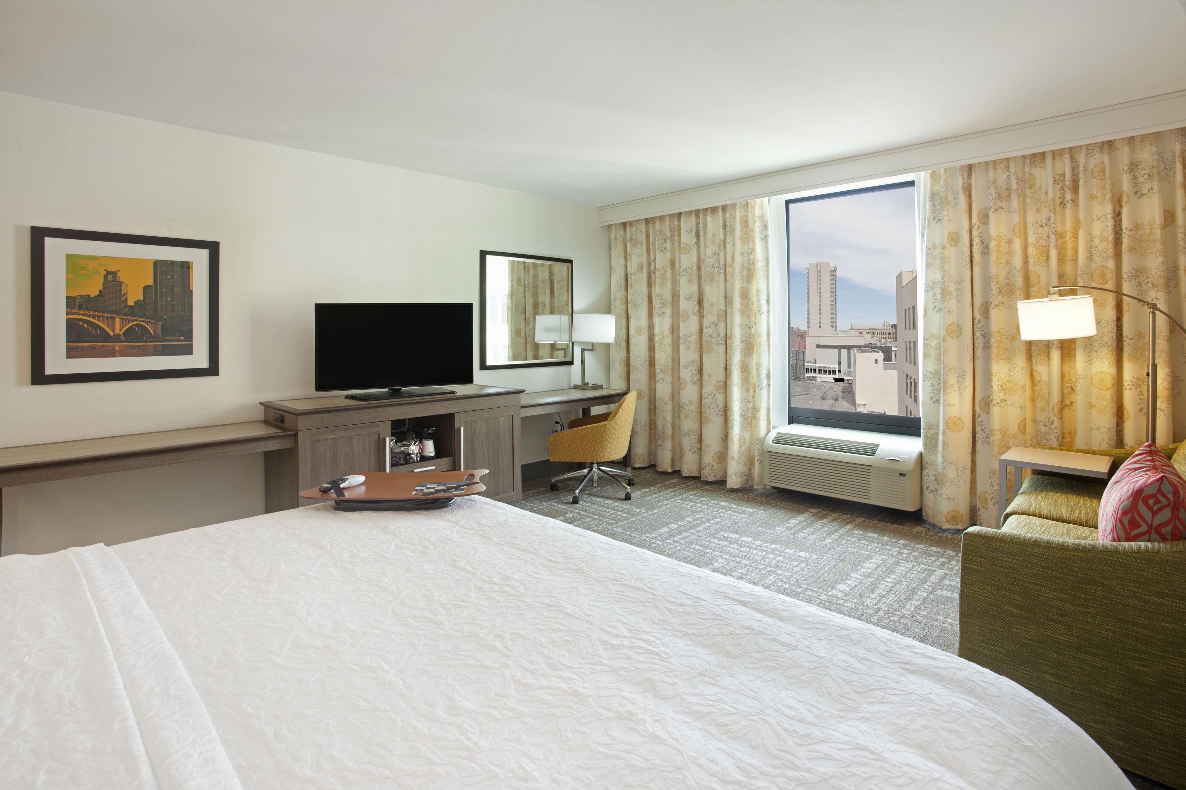 Une chambre d’hôtel équipée d’un lit, d’un bureau, d’une chaise, d’une télévision et d’une grande fenêtre avec des rideaux.