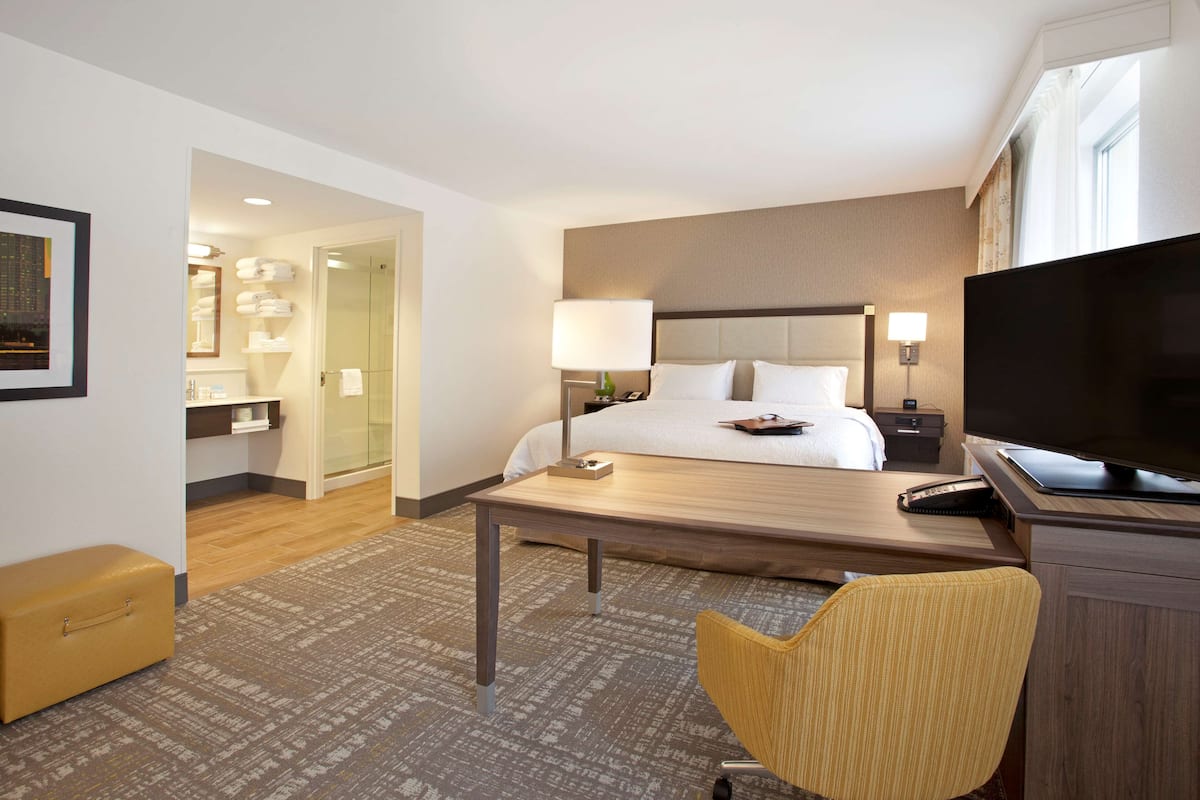 A hotel room with a bed, bedside table, TV, chair, and a desk with a lamp.