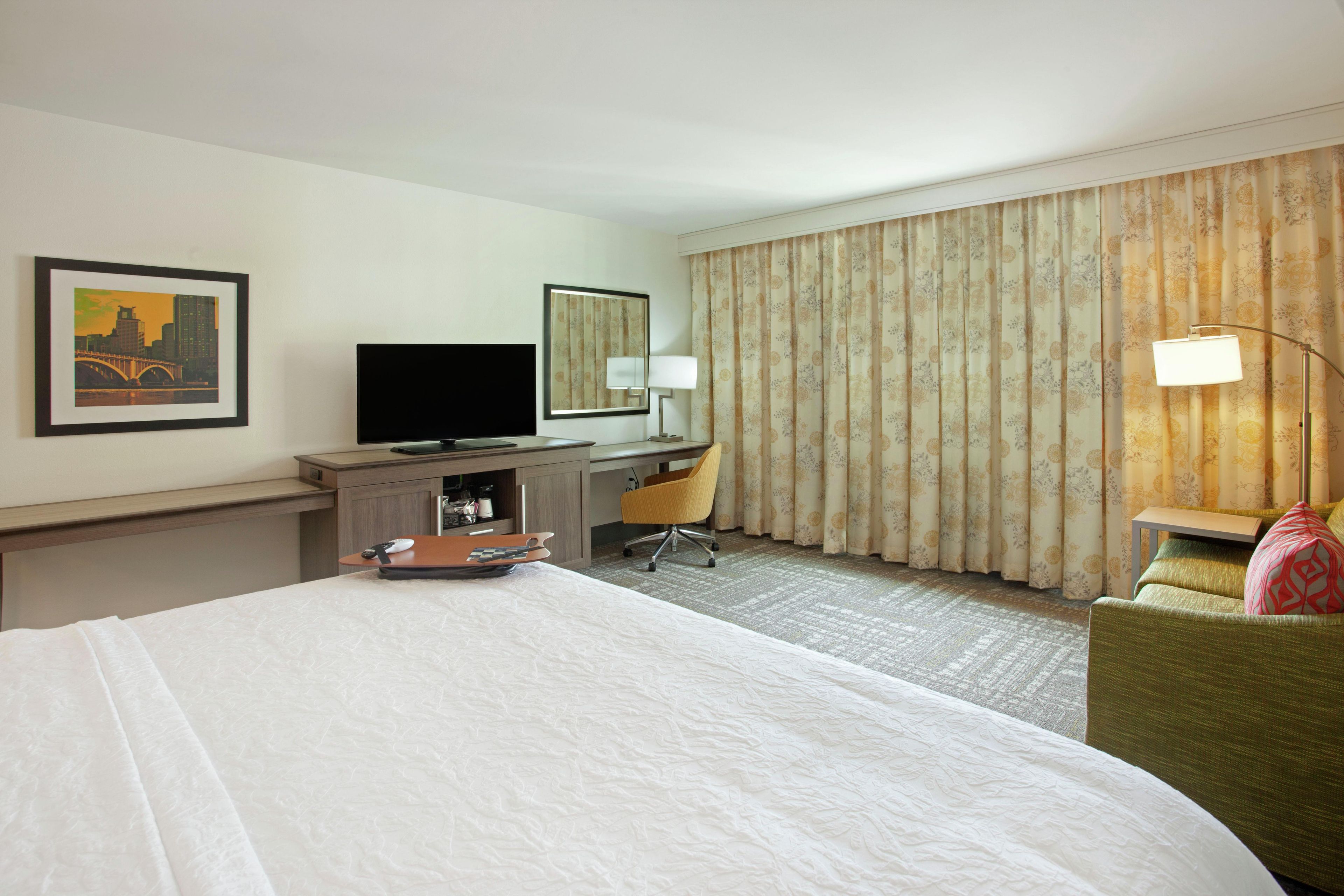 Een hotelkamer met een bed, een bureau met een televisie, een stoel, een lamp en een groot raam met gordijnen.
