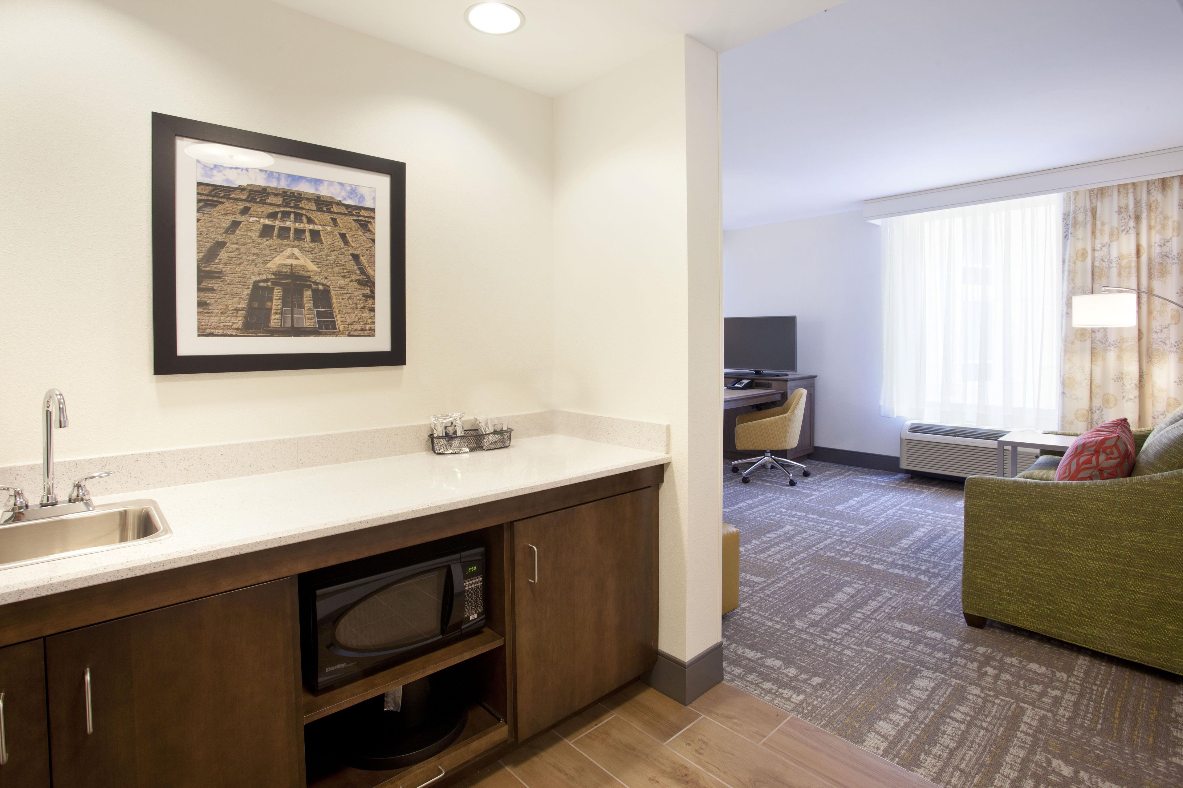 Une chambre d’hôtel avec une kitchenette, un bureau équipé d’un ordinateur, un canapé vert et une grande fenêtre avec des rideaux.