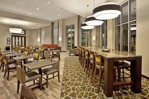 A hotel dining area with multiple tables and chairs, a patterned carpet, and large windows. - Hampton Inn & Suites Minneapolis / Downtown (Minneapolis)