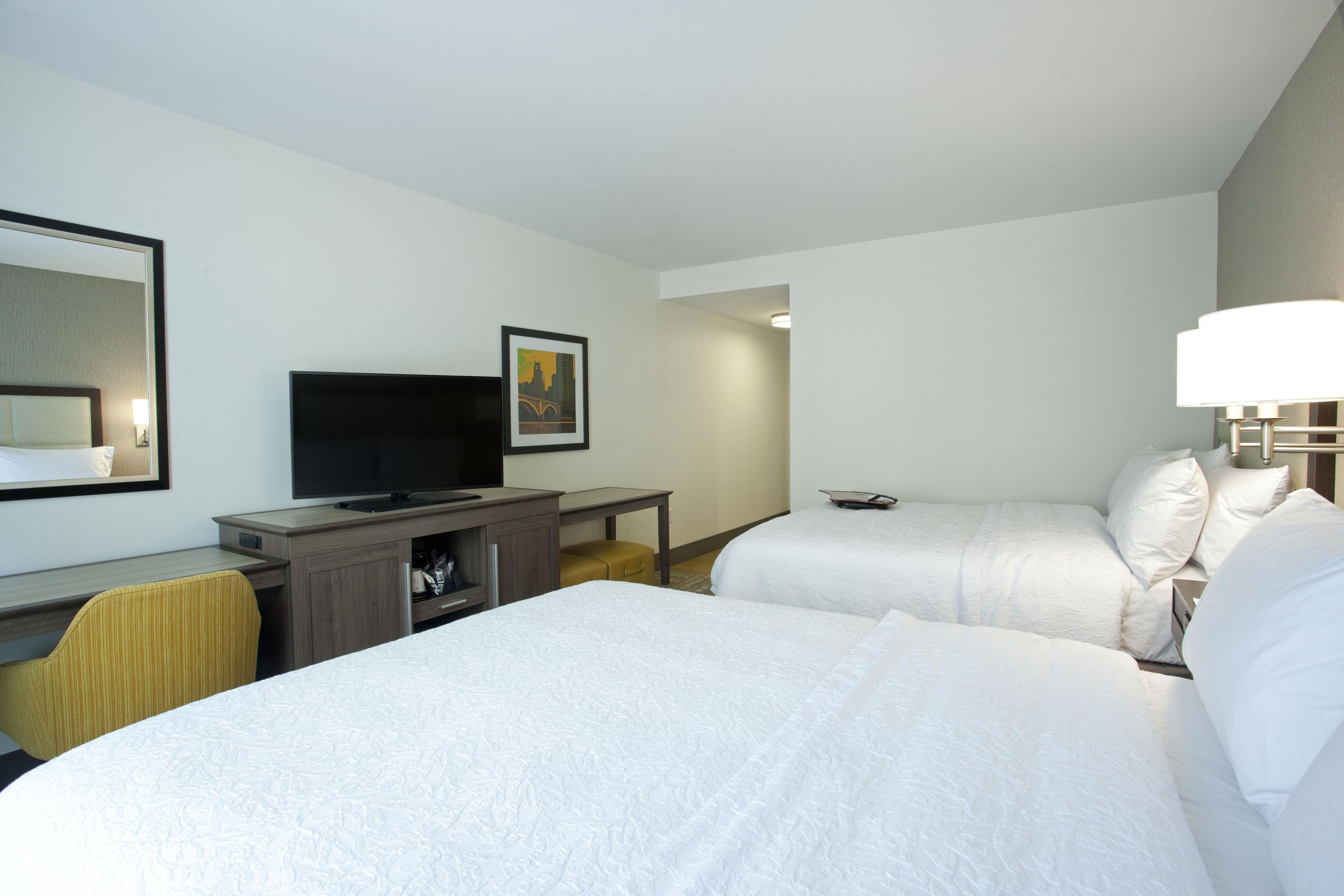 A hotel room with two beds, a desk with a television, a mirror, and a framed picture on the wall.