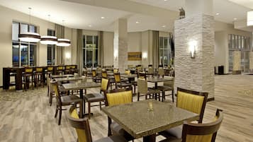 A hotel dining area with multiple tables and chairs, large windows, and a stone accent wall.