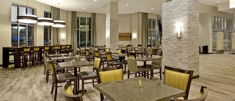 A hotel dining area with multiple tables and chairs, large windows, and a stone accent wall.