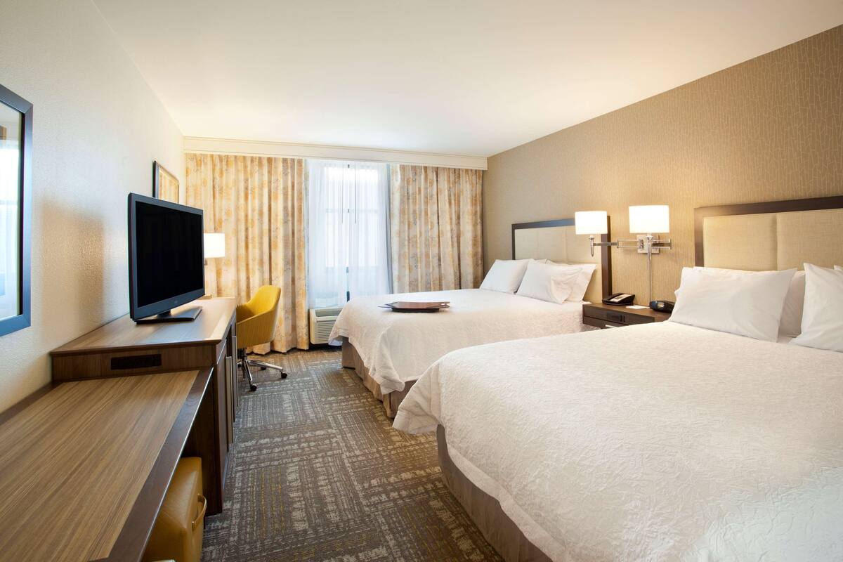 A hotel room with two beds, a desk with a television, a chair, and a window with curtains.