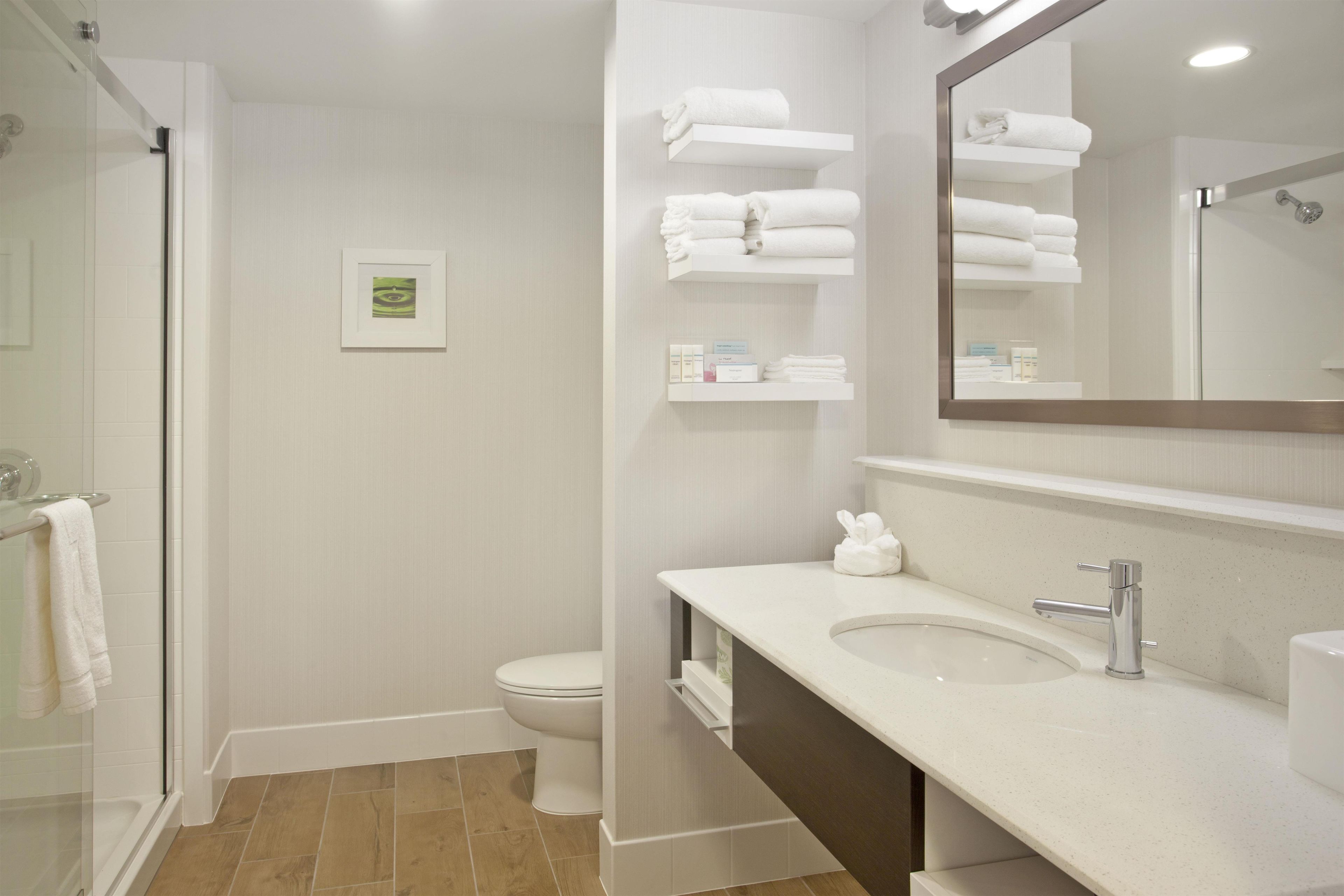 Une salle de bain moderne avec une douche à parois de verre, des toilettes, un lavabo avec un miroir et des étagères contenant des serviettes et des articles de toilette.