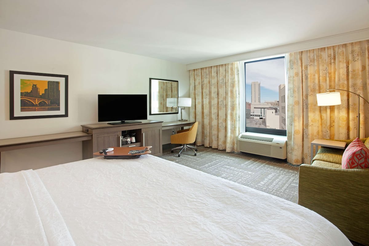A hotel room with a large bed, a desk with a TV, a chair, a window with curtains, and a city view.
