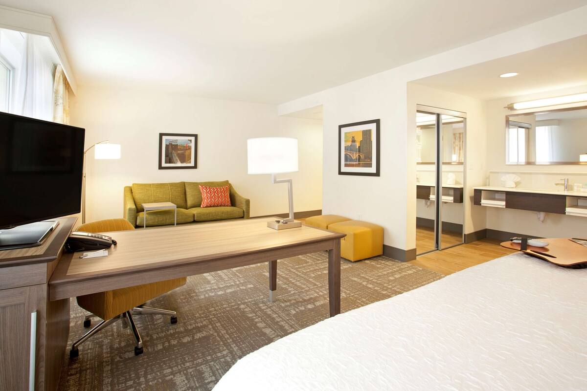 A hotel room with a bed, a desk, a sofa, a television, a mirror, and a bathroom visible through an open door.