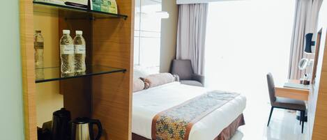 Una habitación de hotel con cama, escritorio, silla, estante con artículos de aseo y ventana con cortinas.