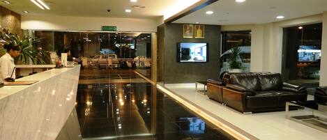 A modern hotel lobby with a reception desk, seating area, and a television screen.