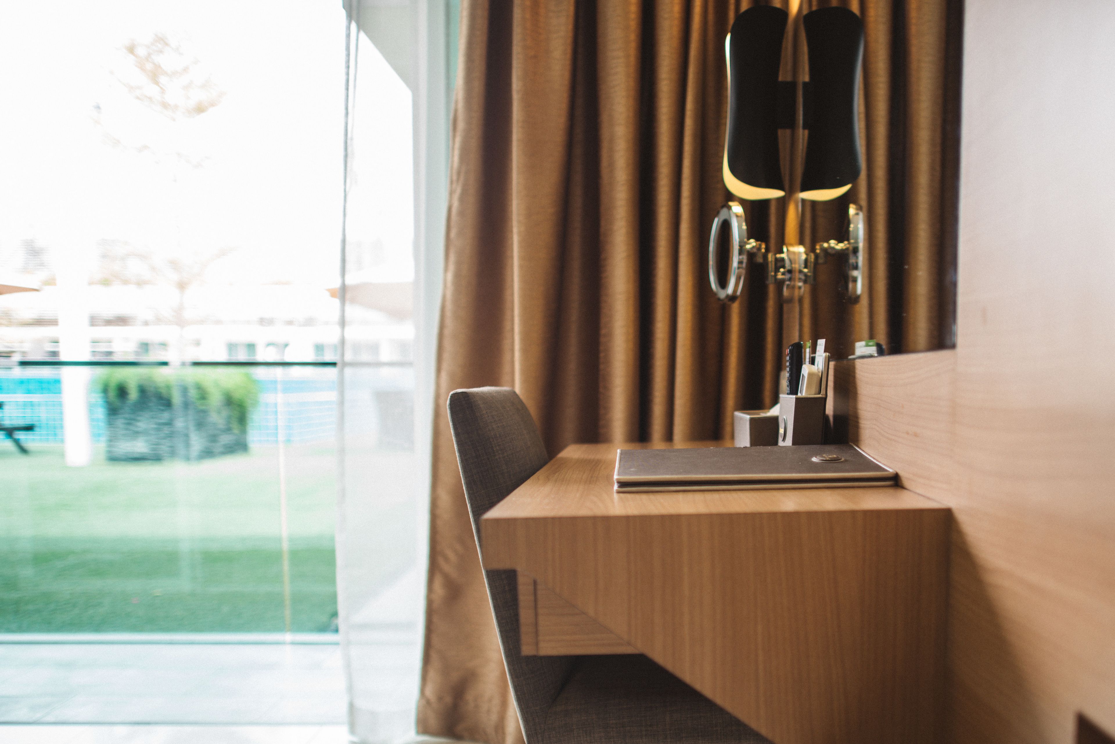 A hotel room with a wooden desk, a laptop, and a lamp. There is a window with curtains and a view of a garden.