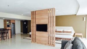 Una habitación de hotel moderna con un televisor de pantalla plana montado en un panel de madera, una cama con colcha color beige y marrón, un sofá gris y un taburete de madera pequeño.