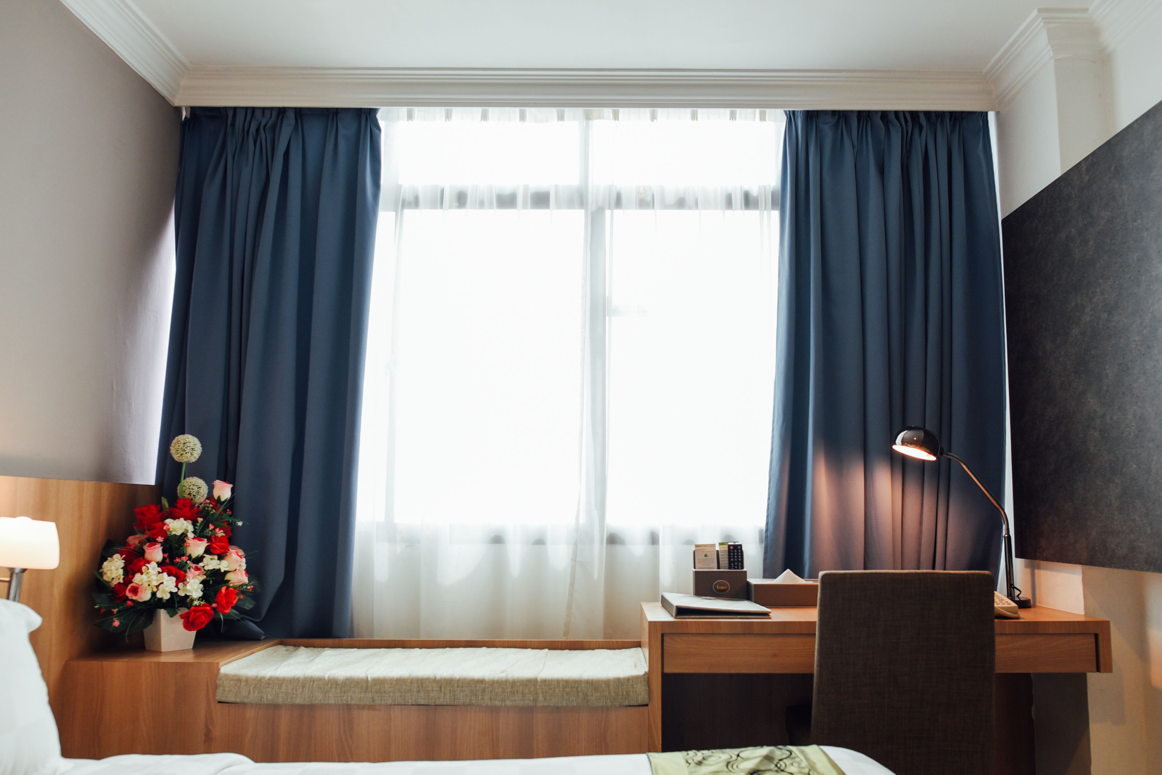 A hotel room with a bed, a desk, a chair, a lamp, and a window with curtains.