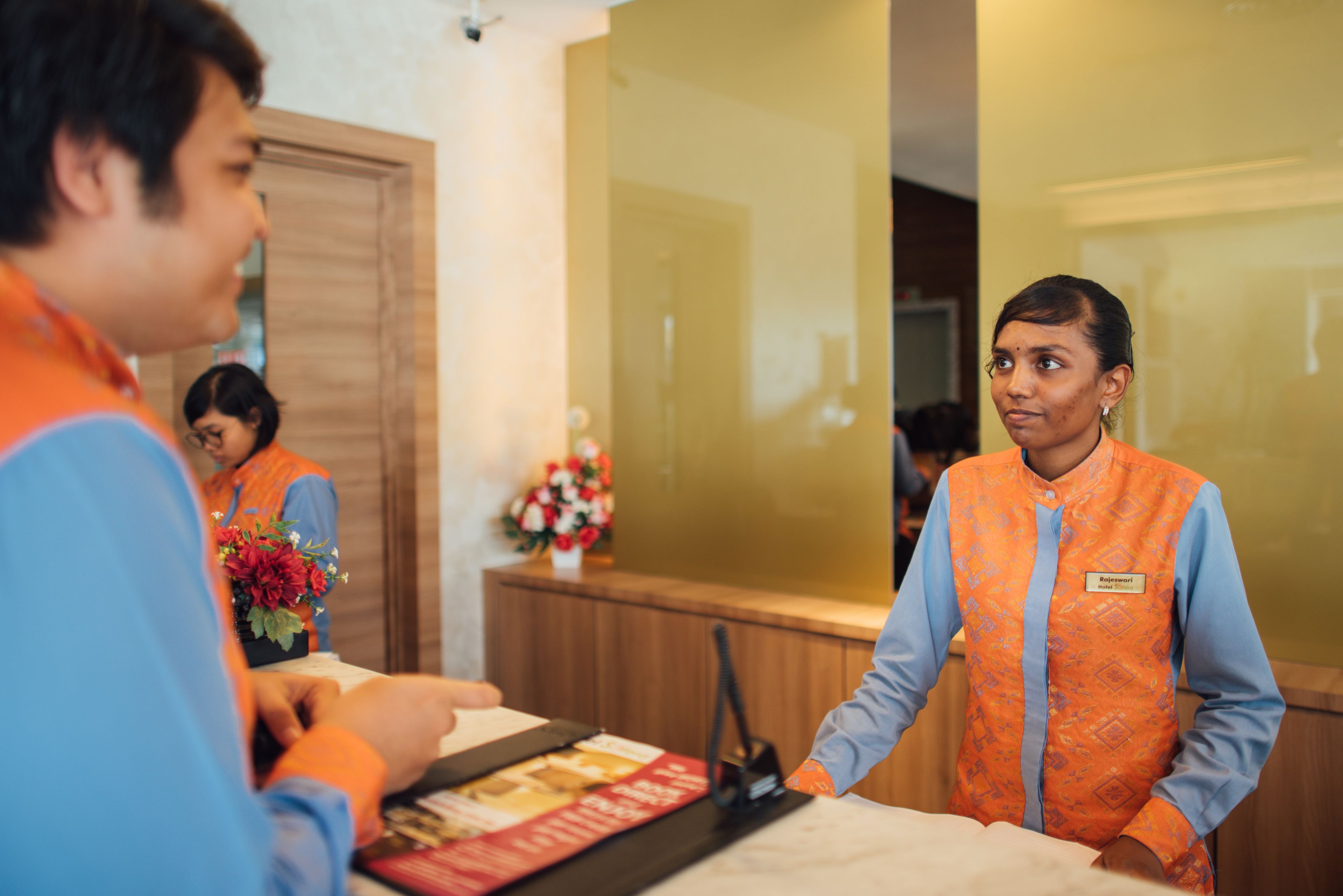 A hotel reception area with a wooden reception desk, a name tag on the staff member, and a flower arrangement on the counter.