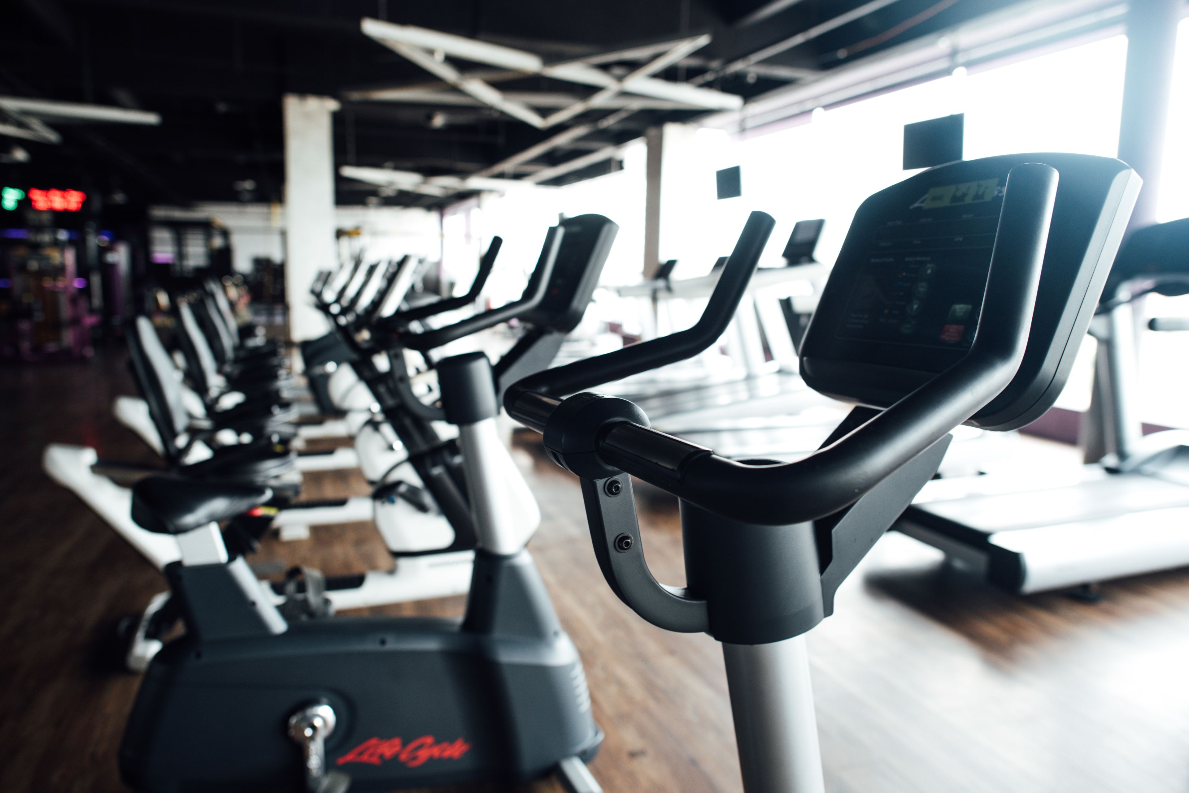 A row of stationary bikes in a gym with a wooden floor and a bright window in the background.