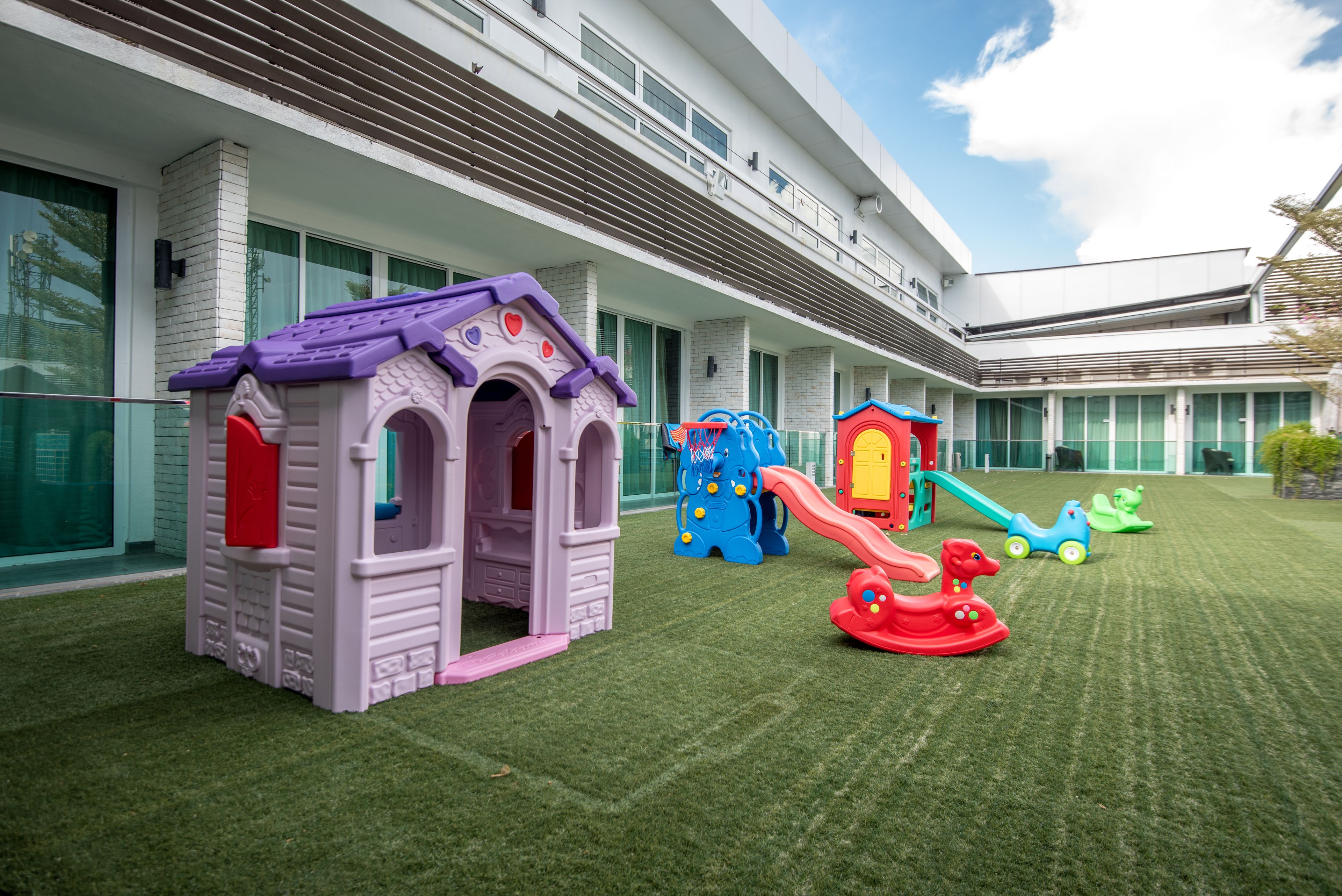 Zona infantil con diversas estructuras de juego, incluyendo una casita, un tobogán y un coche de juguete, frente a un moderno edificio de hotel.