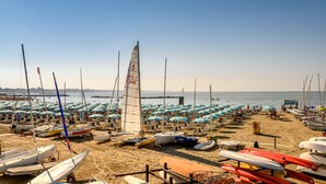 A beach with numerous sailboats and a large number of beach umbrellas.