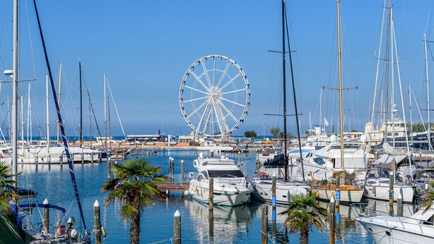 A marina with multiple boats docked, a large Ferris wheel in the background, and a clear blue sky.