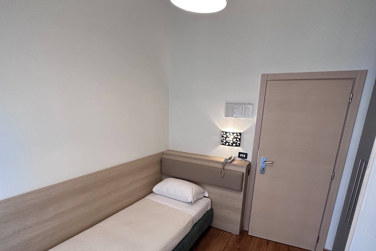 A small, single-bed room with a wooden headboard, a bedside table with a lamp, and a door.