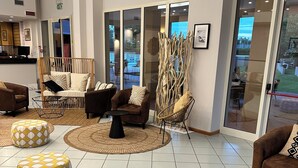 A spacious lobby with a modern design, featuring a mix of seating options including sofas, armchairs, and ottomans. There is a large window providing a view of the outdoors, and the area is well-lit with natural and artificial lighting.