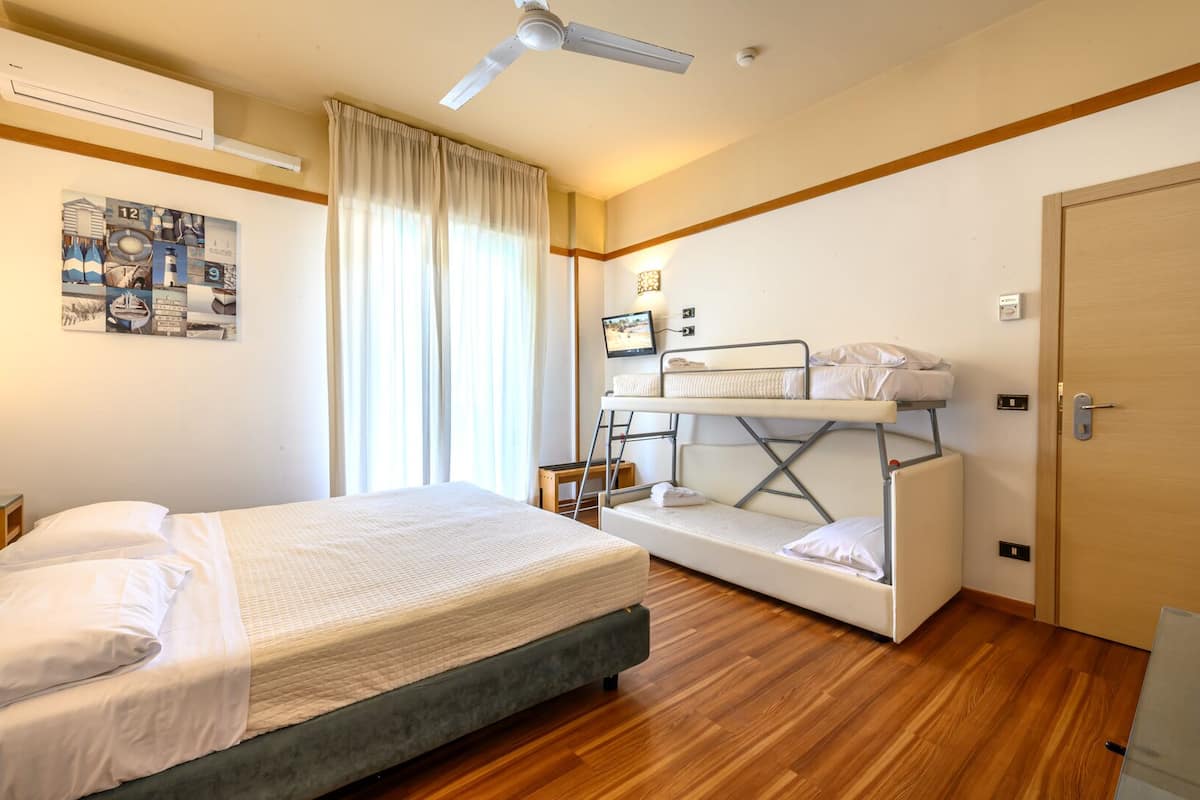 A hotel room with a bed, a bunk bed, wooden flooring, a ceiling fan, and a wall-mounted TV.