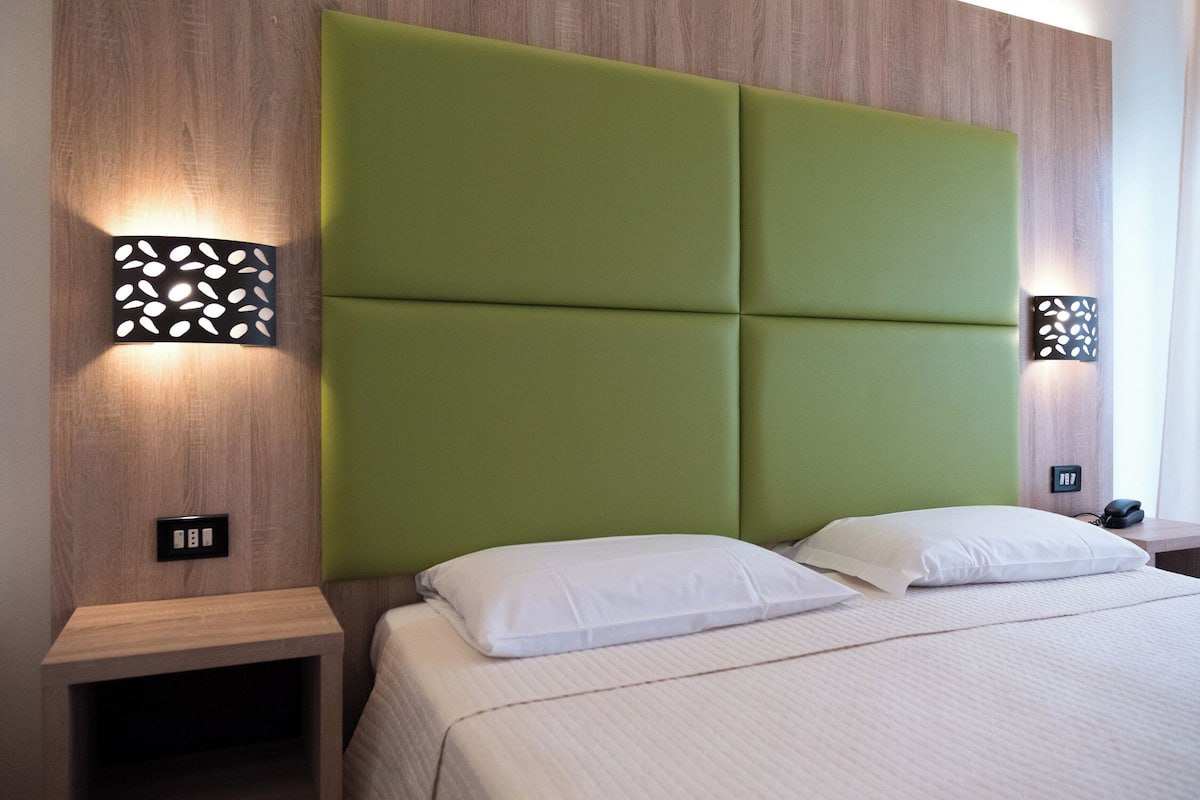 A hotel room with a green padded headboard, two bedside tables, and a bed with white bedding.