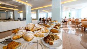 A buffet with various pastries and bread, including croissants and a cake, in a well-lit indoor setting with tables and chairs.